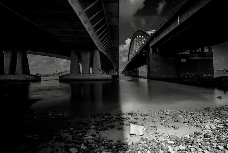 Black-and-white photograph from beneath two parallel bridges, river and stones in the foreground, light between them